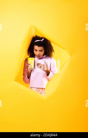 surprised african american woman holding disposable cup and looking down through hole in ripped paper on yellow background Stock Photo