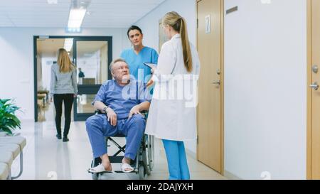 In the Hospital Hallway, Nurse Pushes Elderly Patient in the Wheelchair, Doctor Talks to Them while Using Tablet Computer. Clean, New Hospital with Stock Photo