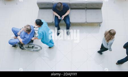 High Angle Shot in the Hospital Lobby, Young Man Waits for Results while Sitting and Using Mobile Phone, Doctors, Nurses and Patients Walk Past Him Stock Photo