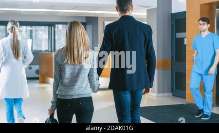 In the Hospital Young Couple Walks to Reception. Medical Personnel, Doctors, Nurses, Assistants and Patients in the Busy Lobby. New, Modern Medical Stock Photo
