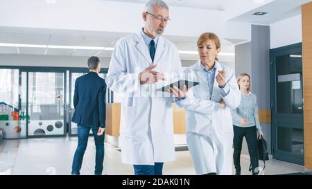 In the Hospital, Busy Doctors Talk, Using Tablet Computer While Walking Through the Building. New Modern Fully Functional Medical Facility. Stock Photo