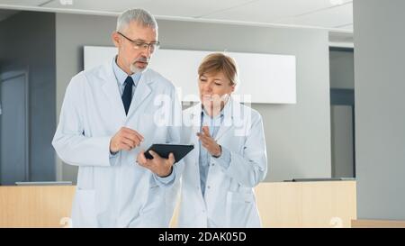 In the Hospital Doctors have Discussion while Using Tablet Computer. They're Walking through the Hallway. In the Background Patients and Medical Stock Photo