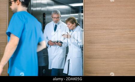 In the Hospital Two Doctors Walk out of the Elevator while have Discussion while Using Tablet Computer. In the Background Patients and Medical Stock Photo