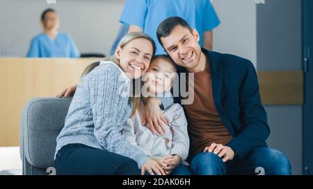 Portrait of the Young Happy Family in the Hospital. Handsome Father, Beautiful Mother and Cute Little Daughter Sitting in the Lobby of the Medical Stock Photo