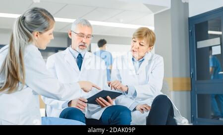 In the Lobby of the Hospital Three Professional Doctor Have Discussion while Sitting. They Use Tablet Computer. Busy Modern Hospital with Best Stock Photo