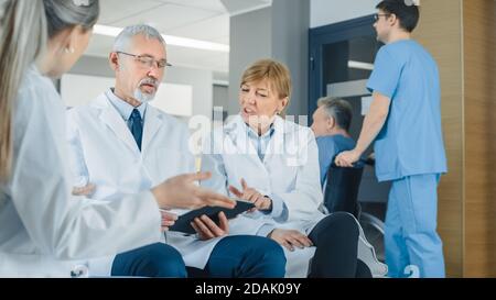 In the Lobby of the Hospital Three Professional Doctor Have Discussion while Sitting. They Use Tablet Computer. Busy Modern Hospital with Best Stock Photo