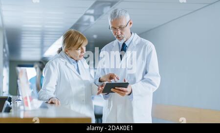 In the Hospital Two Doctors Standing at the Reception Desk, Talking and Sharing Information on Tablet Computer. Modern Hospital with Patients and Stock Photo