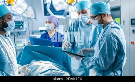 Professional Surgeons and Assistants Talk and Use Digital Tablet Computer During Surgery. They Work in the Modern Hospital Operating Room. Stock Photo