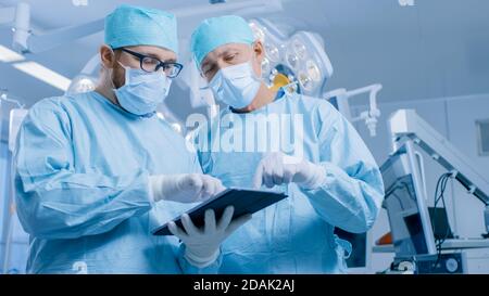 Two Professional Surgeons Use Digital Tablet Computer while Standing in the Modern Hospital Operating Room. Stock Photo