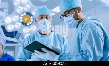 Two Professional Surgeons Use Digital Tablet Computer while Standing in the Modern Hospital Operating Room. Stock Photo