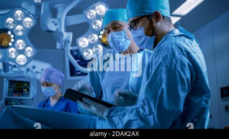 Professional Surgeons and Assistants Talk and Use Digital Tablet Computer while Standing in the Modern Hospital Operating Room. Stock Photo