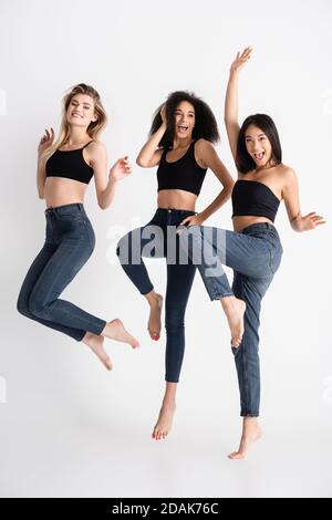 excited interracial women in denim jeans jumping and posing on white Stock Photo