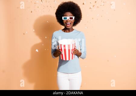 Photo portrait of curly excited amazed dark skinned girl keeping box with popcorn flying in air watching movie wearing 3d glasses isolated on beige Stock Photo