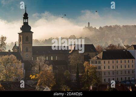 The tower of the church, likely part of St. Michael's monastery church, rises above Bamberg's autumn rooftops, Bavaria, Germany; to the right, hazy in the distance, the tower of Altenburg castle stands on a wooded hill. The proceeds from the historic vineyard at the foot of the Michelsberg, revived in 2009, fund the restoration of the former Benedictine abbey of St. Michael. Altenburg was the residence of the Bamberg prince-bishops from 1305 to 1553. Stock Photo