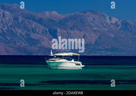 Small modern boat on the clear Aegean Sea with a towering mountainous backdrop (Crete, Greece) Stock Photo