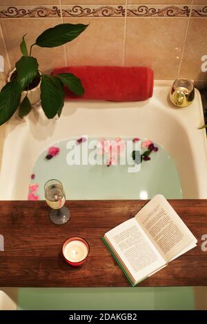 Time for Yourself. Relax at home. Bath tub with flower petals. Book, candles and glass of wine on a wood tray. Organic Spa Relaxation in comfort cozy Stock Photo