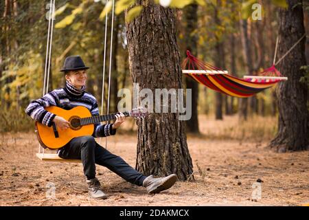 young handsome attractive man in sweater playing guitar in forest sitting on swing with hammock on background. Casual man, life style.