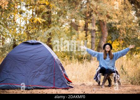 Young woman tourist sitting near tent in blue sweater and gray hat and enjoy freedom at autumn forest sunset Stock Photo