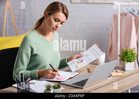 Freelancer writing on notebook while holding document with charts near laptop on table Stock Photo