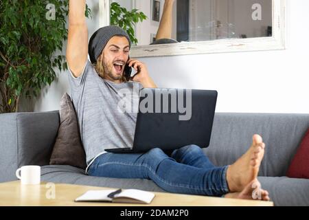 Handsome young business caucasian man working at home using laptop and talking with mobile phone feeling happy Stock Photo