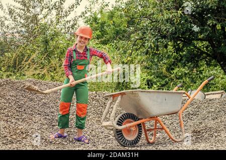 game of builder and construction. Outdoor activity concept. girl who is transporting rubble in a wheelbarrow. construction work. girl builder takes a shovel of rubble. hardworking youth. Stock Photo