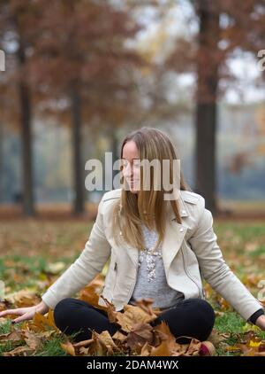 Young Blonde Woman Sitting in Autumn Park with Brown Maple Leaves Stock Photo