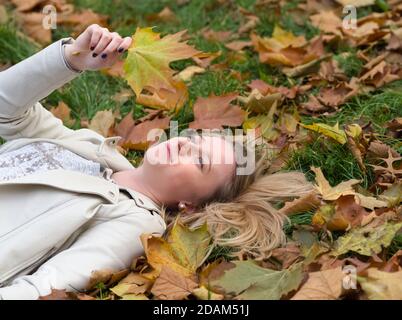 Young Beautiful Blonde Woman Laying in the Grass in Autumn Park among Maple Leaves Stock Photo