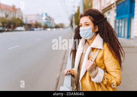 Beautiful afro haired woman wearing protective medical face mask stand on the street of city. Social distancing, quarantine. Stock Photo