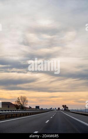A motorway straight that gets lost against a cloudy sunrise sky in vertical format, a car overtakes a truck further down the road. Stock Photo