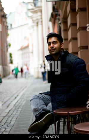 A south asian Indian ethnicity man sitting in an open street side restaurant located in central europe. Low angle shot of a smiling man looking at cam Stock Photo