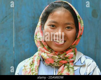 Pretty Northeast Indian Apatani pre-teen girl wears a colorful head scarf and smiles for the camera in front of blue wooden door background.