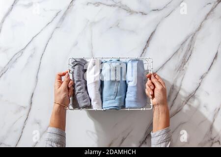 Neatly folded clothes and pyjamas in the metal mesh organizer basket on white marble table. Stock Photo