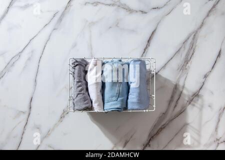 Neatly folded clothes and pyjamas in the metal mesh organizer basket on white marble table. Stock Photo