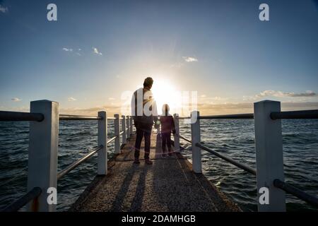 Looking out over a pier against the ocean with a father and son in silhouette, a dusk horizon sky Stock Photo