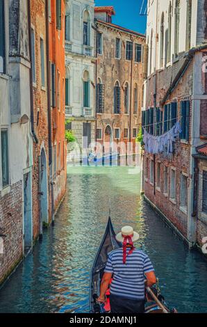 Gondola sailing narrow canal in Venice between old buildings with brick walls. Gondolier dressed traditional white and blue striped short-sleeved polo Stock Photo