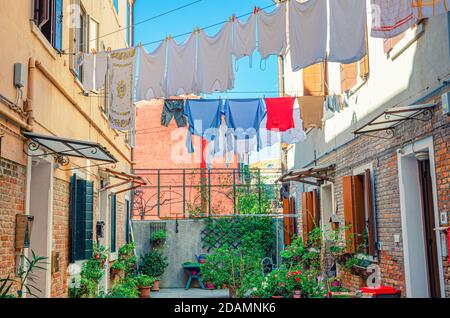 Typical italian courtyard between buildings with brick walls, green plants and flowers in flowerpot, small tables, wet clothes hanging on cord, Murano Stock Photo
