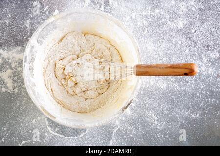 Close-up glass bowl with dough. Beat a delicious dough with flour, eggs, milk and sugar. The dough is ready to cook. Glass table. Sprinkle with flour. Stock Photo