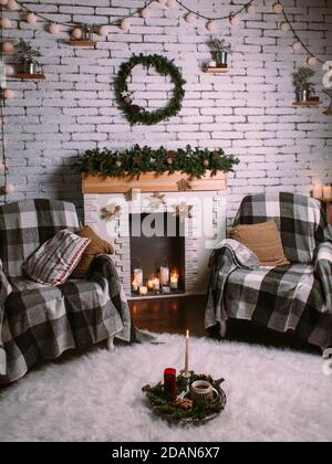Decorative fireplace decorated with New Year's garland and burning candles in glass cups. Round green wreath hanging on white brick wall. Cozy Christm Stock Photo
