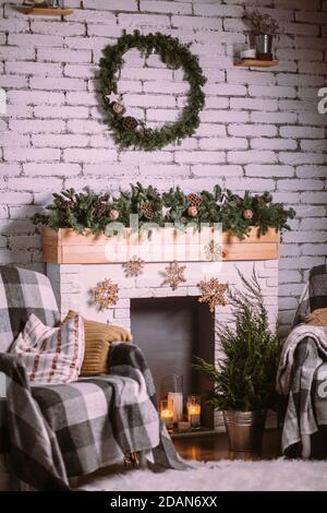 Decorative fireplace decorated with New Year's garland and burning candles in glass cups. Round green wreath hanging on white brick wall. Cozy Christm Stock Photo