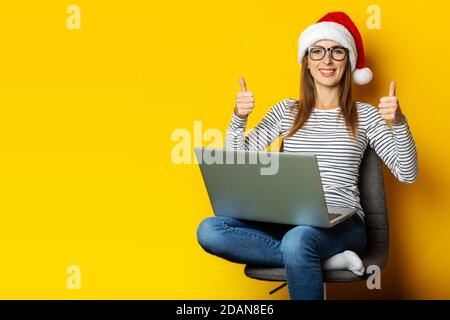 Young woman in a Santa hat with a happy face sits on a chair and holds a laptop and rejoices at something on a yellow background. Banner. Stock Photo