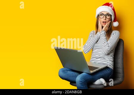 Young woman with a surprised face and a laptop, wearing a Santa hat and sitting on a chair on a yellow background. Banner. Stock Photo