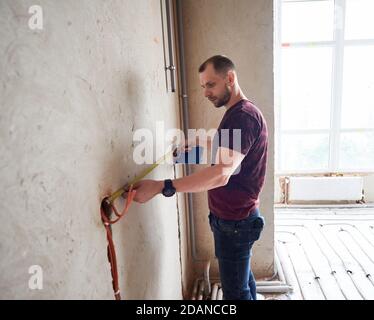 Side view of serious man holding notebook and using measuring tape in room with underfloor heating pipes. Handyman in shirt taking measurements while working on renovation of apartment.