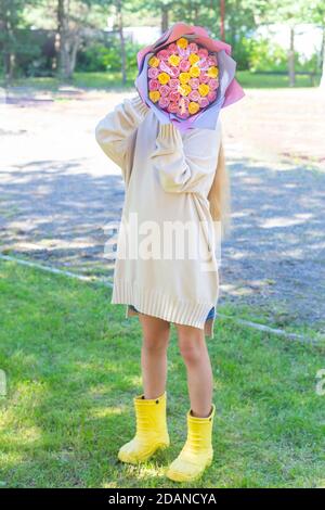 Caucasian girl in a long light sweater and yellow rubber boots stands in nature and covered her face with a bouquet of multi-colored roses. Stock Photo