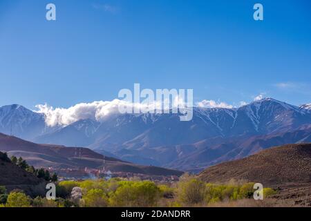 Daytime wide angle shot of Beautiful landscape of snow capped mountains and bushes and a village in the valley. Atlas, Morocco. Stock Photo