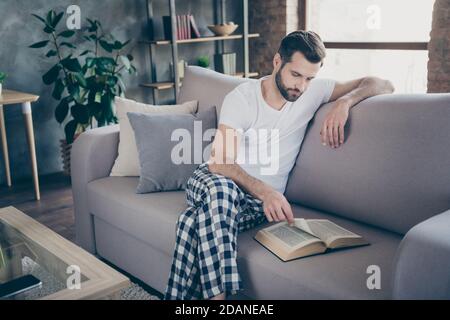 Portrait of his he nice attractive focused bearded guy sitting on divan reading interesting paper book page print spending free time leisure at modern Stock Photo