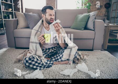 Full length photo of sick ill guy sit carpet hold hot tea beverage mug sneeze napkin cover plaid checkered blanket wear nightwear sleepwear in house Stock Photo