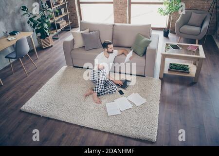 Nice attractive focused guy sitting on carpet studying learning working remotely online analyzing documents at modern loft industrial open space Stock Photo