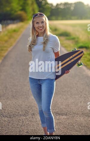Portrait of a fit beautiful middle-aged woman with an active lifestyle smiling and looking at camera while holding a longboard on a sunny road in the Stock Photo
