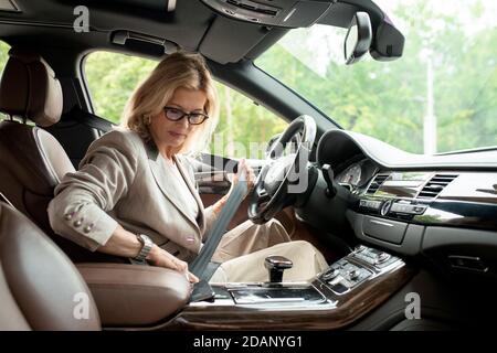 Mature businesswoman in elegant casualwear sitting in car and fastening seatbelt Stock Photo