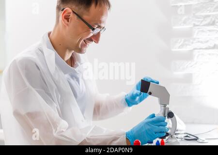 A male doctor or scientist looking through a microscope on a table with laptop computer in background